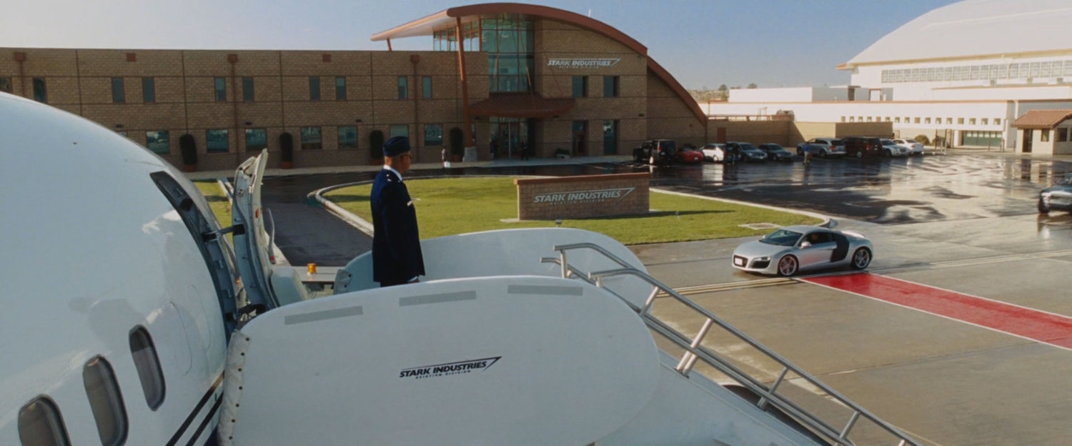 Rhodey stands on a airplane stairway as silver Audi approaches at the Stark aviation airfield.