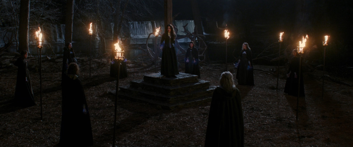 Agnes tied to a post surrounded by witches in small wooded clearing.