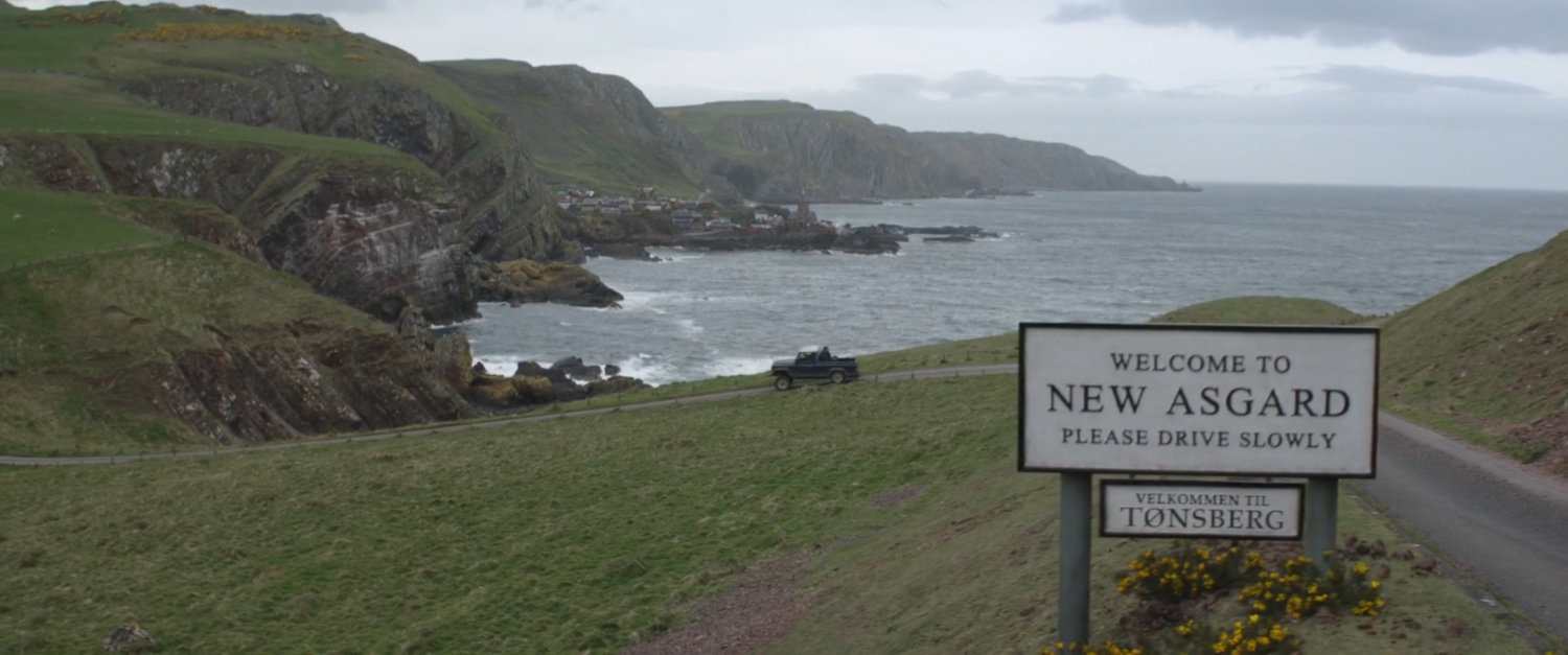 Pickup truck driving down hill by the coast. Sign text: Welcome to New Asgard, please drive slowly.