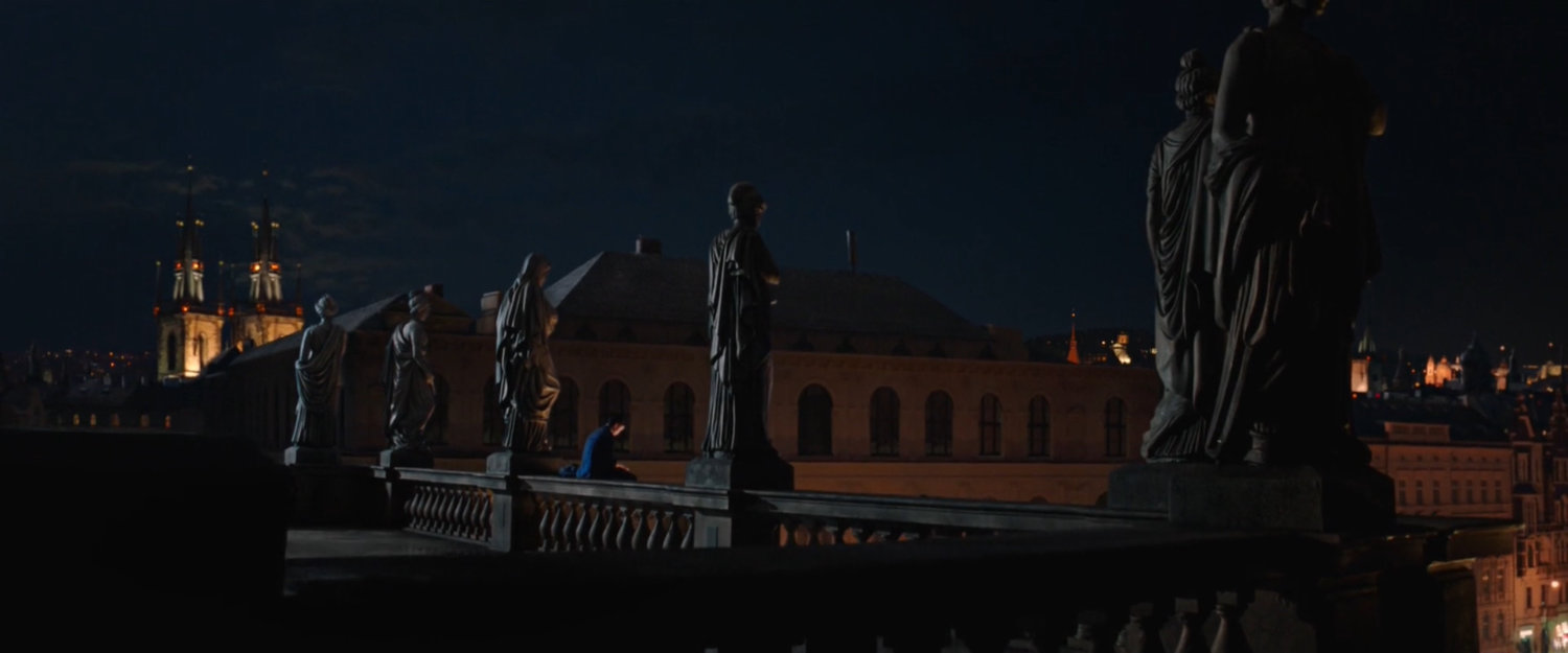 Peter sits on the railing of a concrete balcony in Prague, surrounded by stone statues.