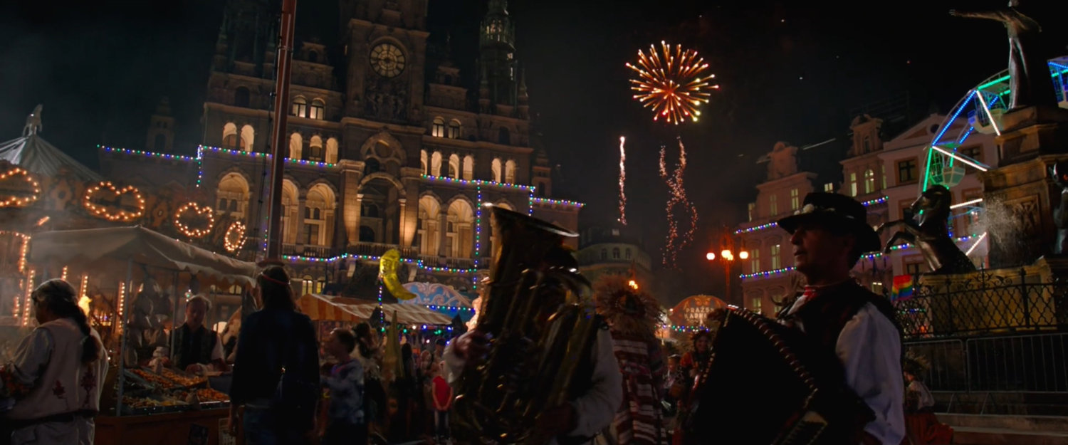 A nightime carnival in a crowded Prague square with fireworks in the distance.