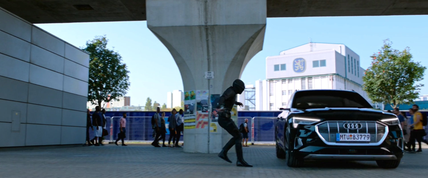 Spider-Man in black outfit approaches car at Berlin train station. License plate: MTU 83779.