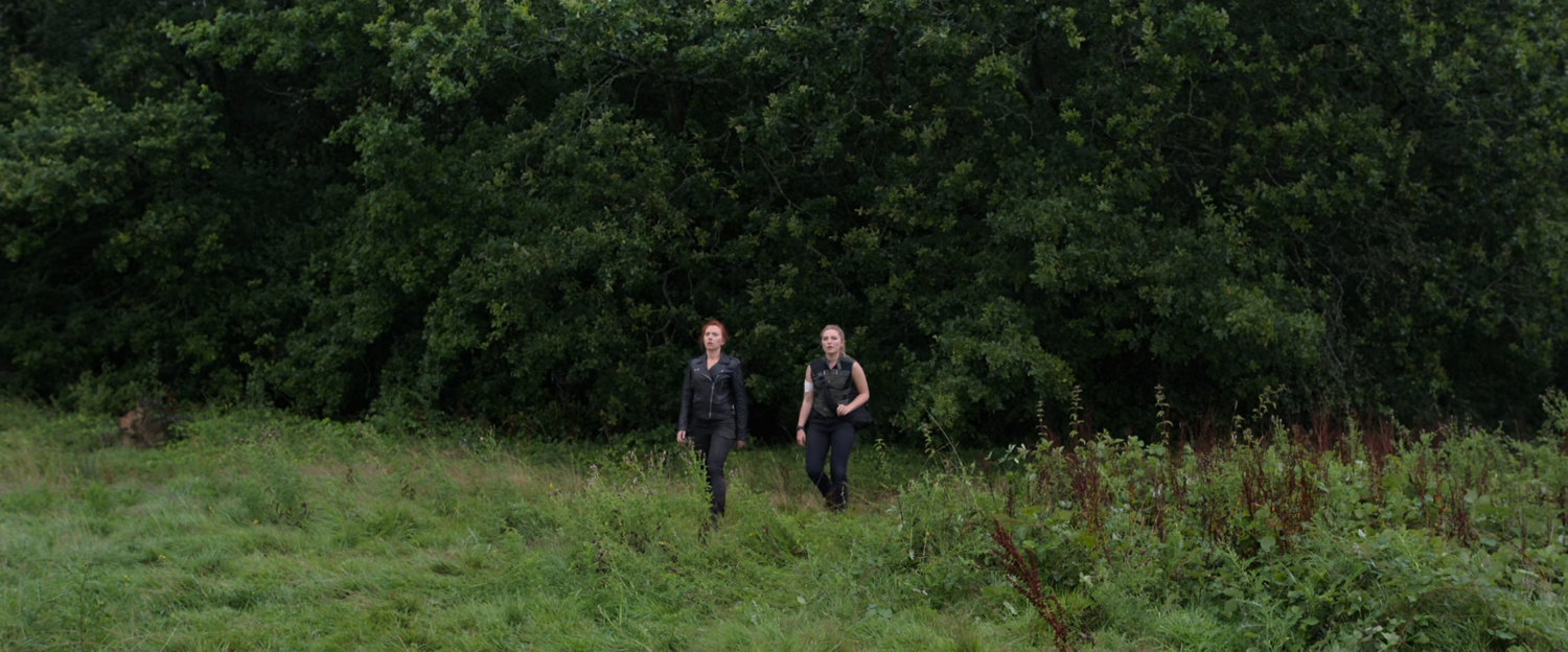 Natasha and Yelena walking in a field near some woods.