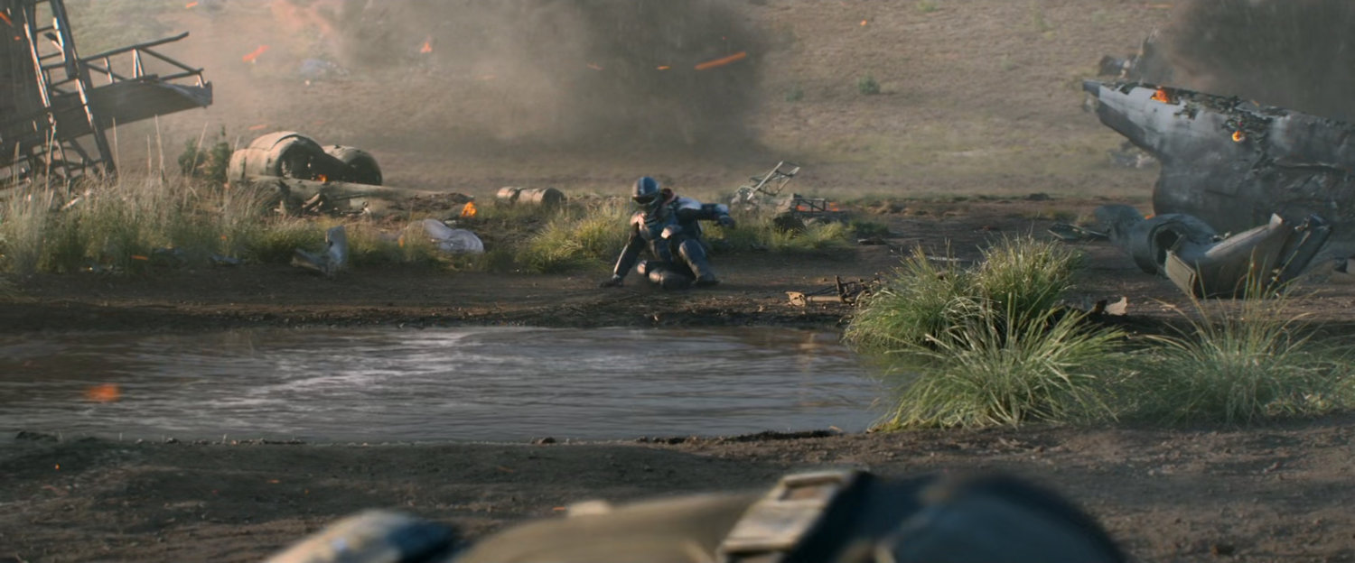 Taskmaster sits by a small pond in a field strewn with debris.