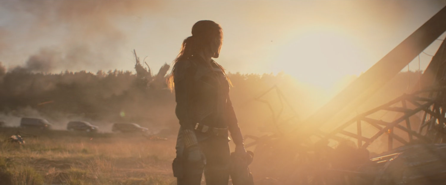 Natasha standing in a field strewn with debris as the sun sets behind her.