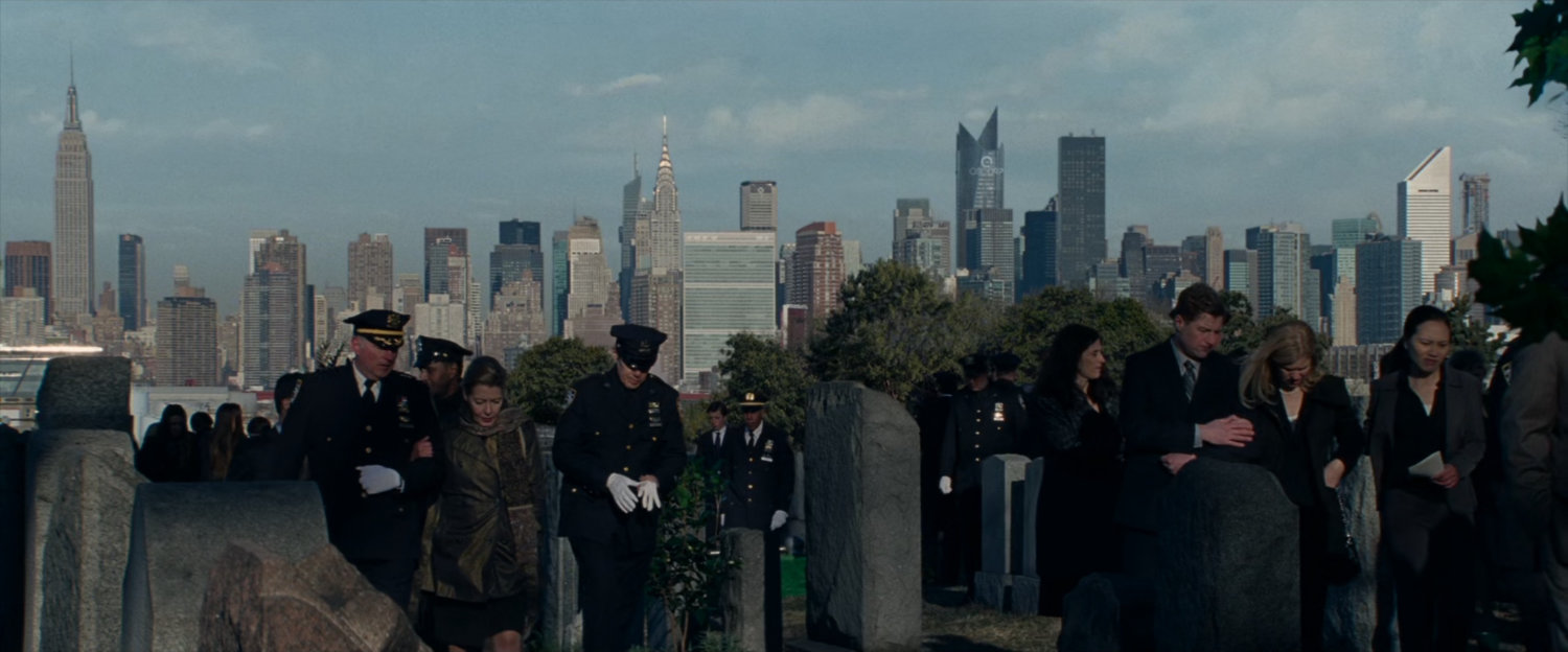 Mourners gather at cemetery with New York in background.