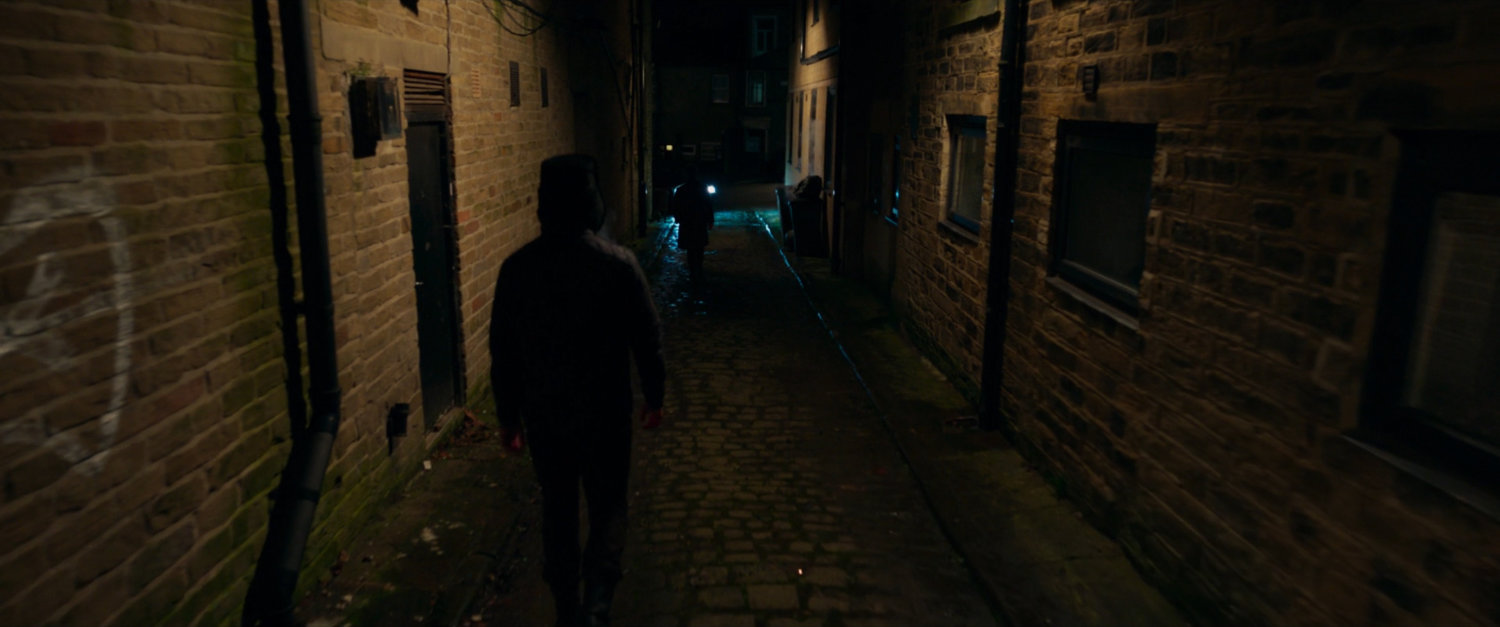 Agent Ross running down a dark alley, followed by another person.
