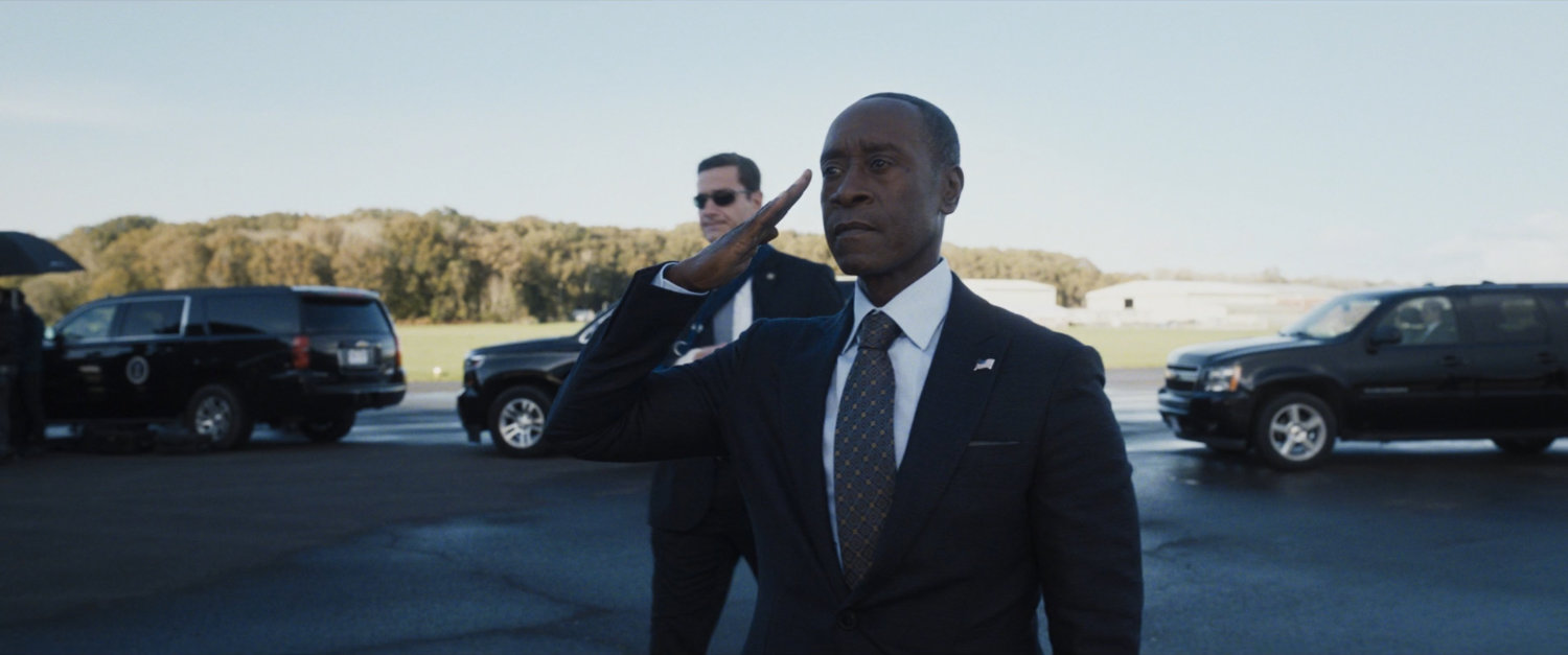 Colonel Rhodes salutes in front of a motorcade on an airfield tarmac.