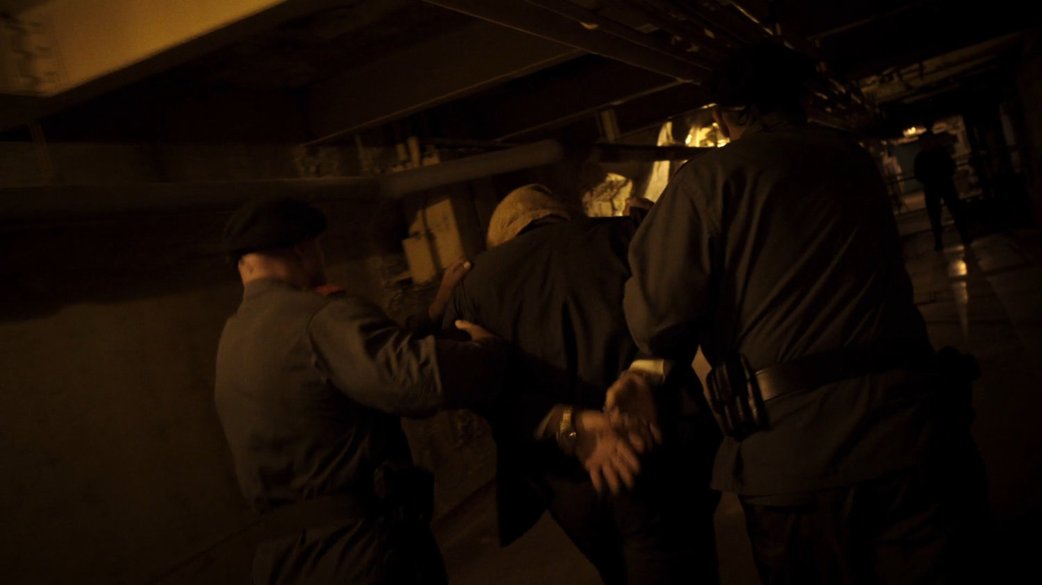 Two guards lead a handcuffed prisoner through a dark underground tunnel.