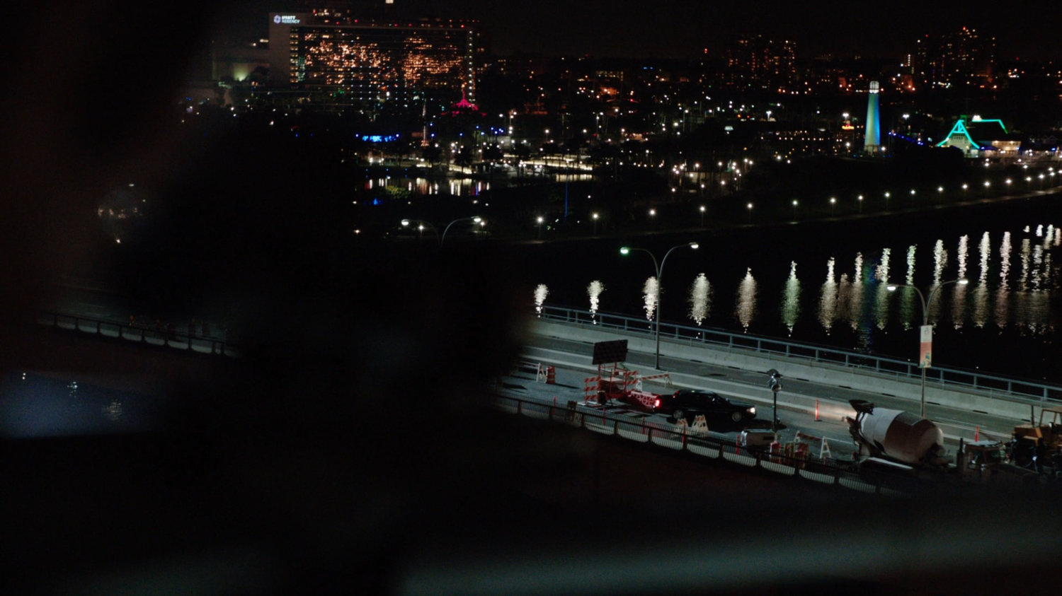 Long high-angle shot of car on bridge by barricades, with city lights in background.