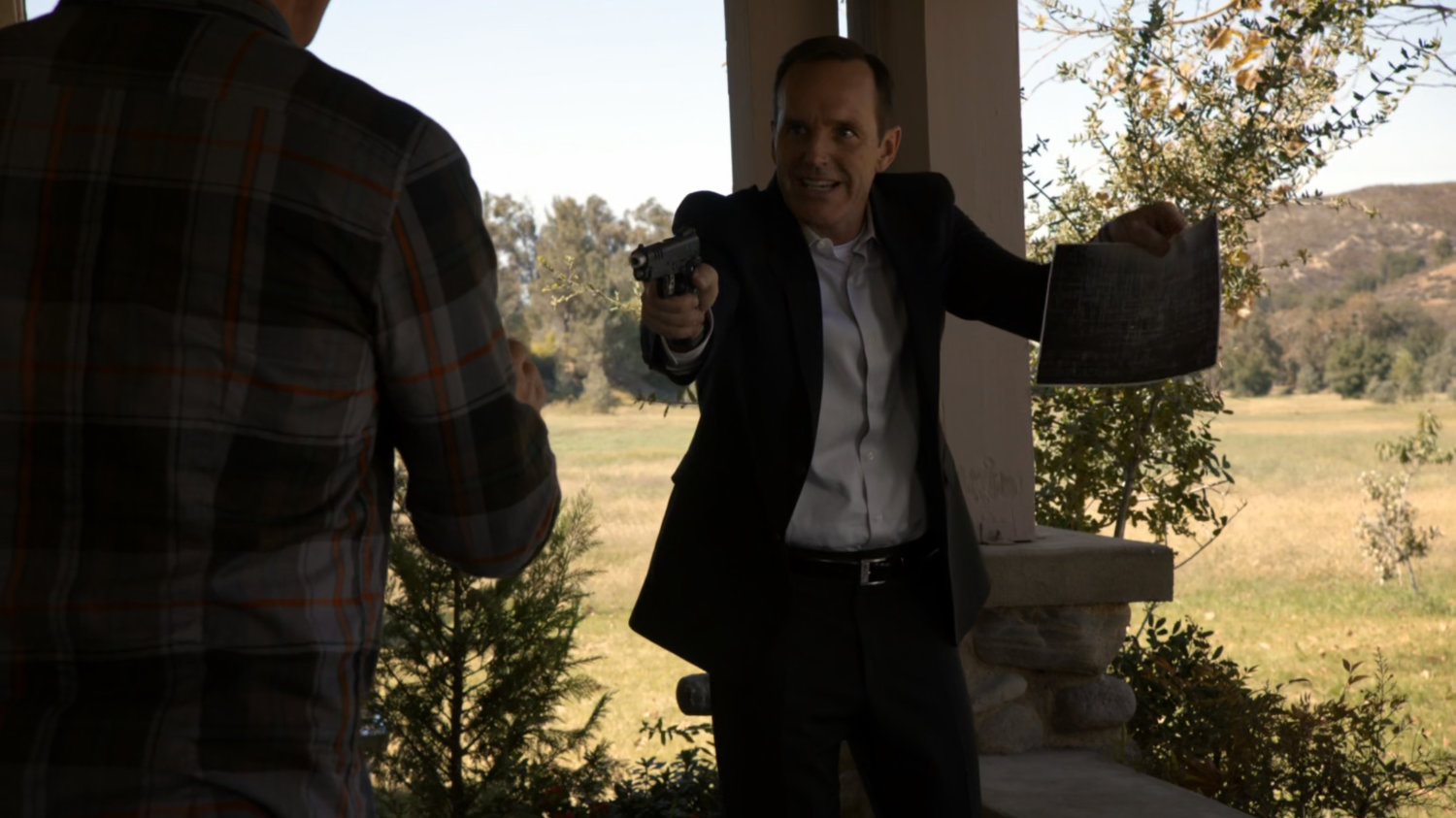 Coulson holds up a photo and a gun to a man on a porch.