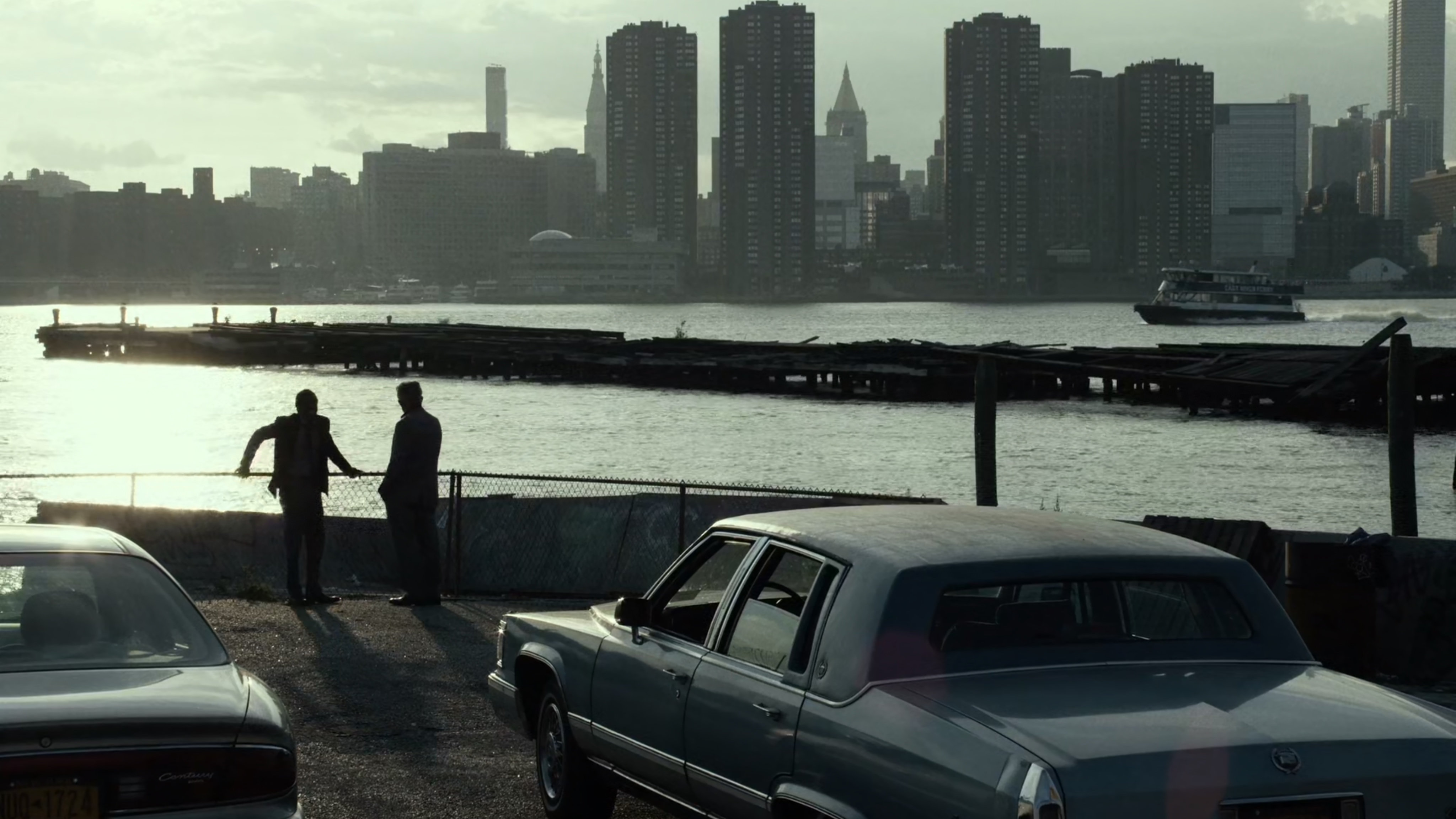 Two men talk in lot by East River with New York City in background.