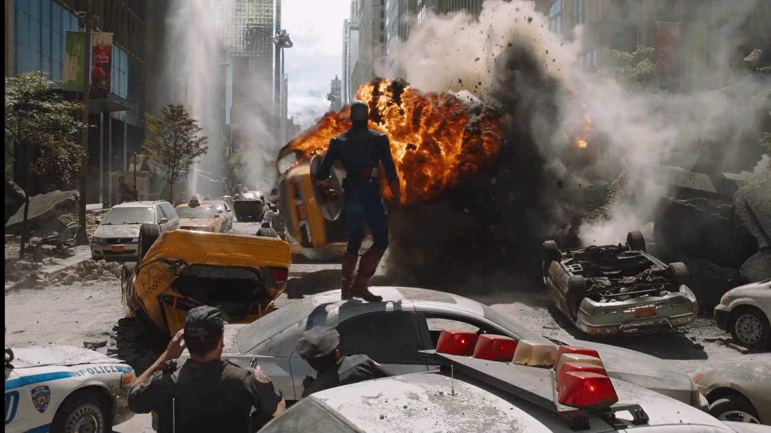 Captain America stands on a wrecked car surveying the explosions and damage.