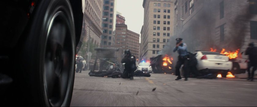 Police shoot at a car, while fires burn behind them.