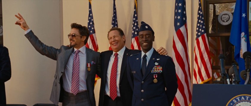 Tony Stark, Senator Stern, and James Rhodes  posing in front of American flags.