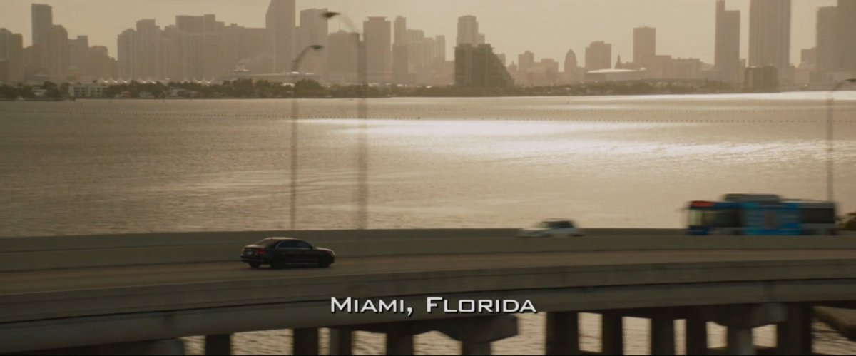 Aerial of car driving over bridge with Miami in background. Text: Miami Florida.