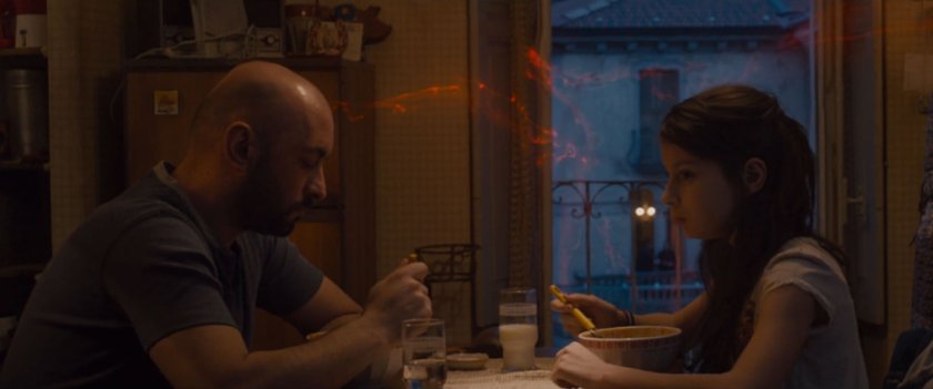 A man and his daughter eat in an apartment with red magic tendrils coming in window.