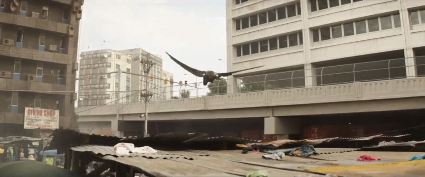 The Falcon flies over marketplace roofs near high rise building.