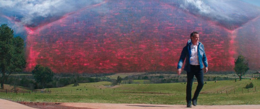 Director Hayward walks away from agreen valley with a red hex in the sky above it.