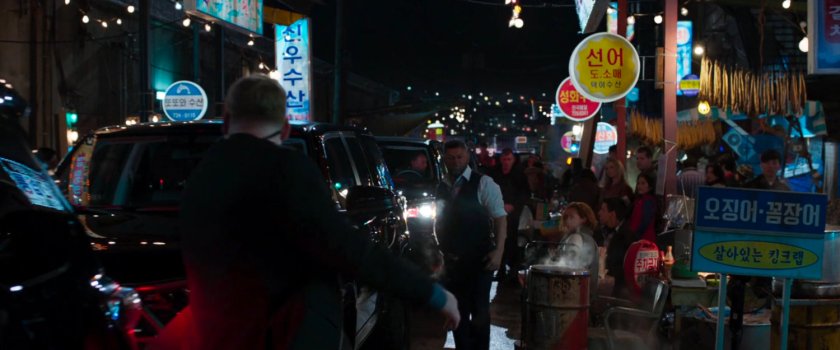 Klaue and a number of black SUVs in a Busan alley/market. 