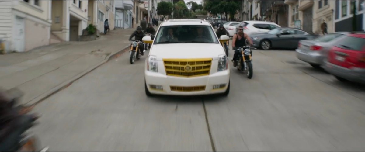 A white SUV flanked by motorcycles drives down a hilly San Francisco street.