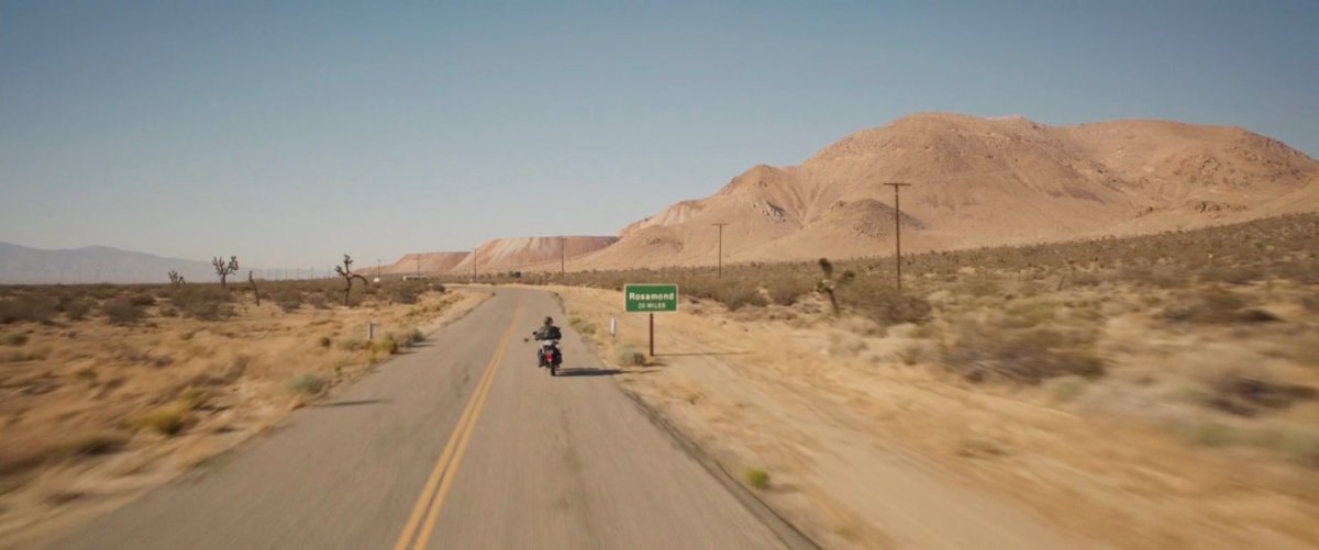 Vers riding motorcycle on desert highway by sign for "Rosamond."
