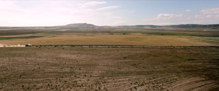 Wide shot of desert plain with car on dusty road.