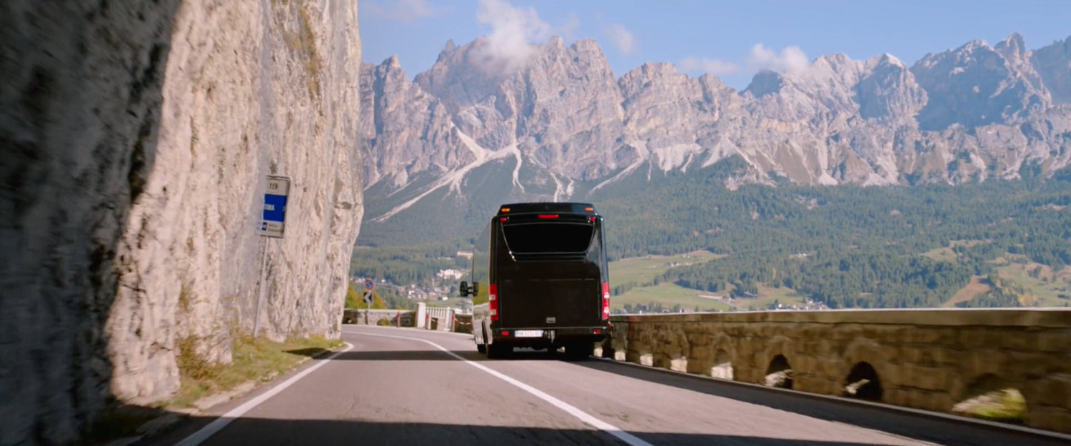 Tour bus on mountain road with giant mountains in background.