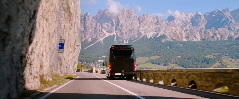 Tour bus on mountain road with giant mountains in background.