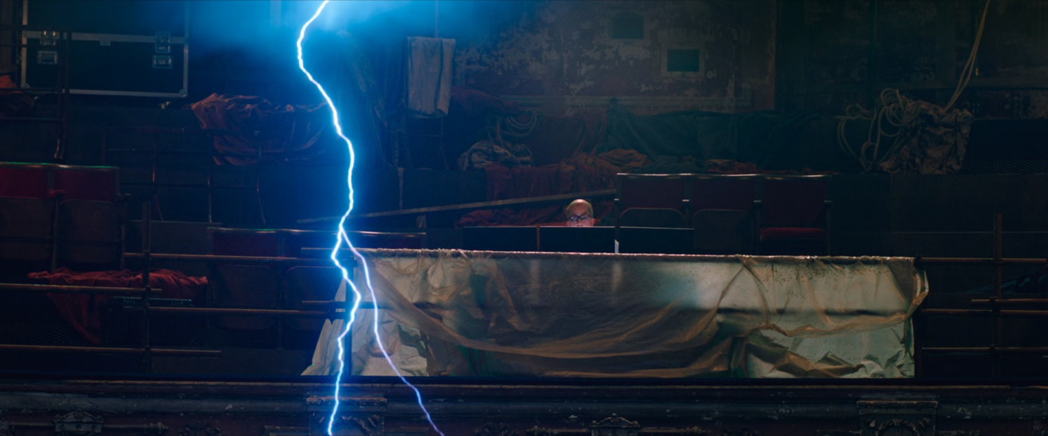 A man sitting in a theater balcony behind computer monitors with a lightning strike near him.