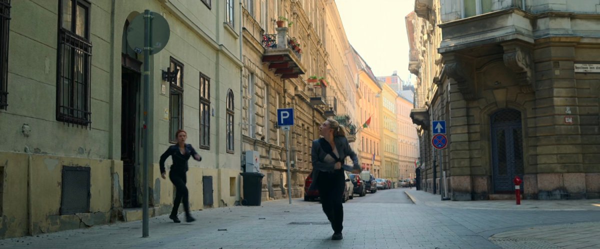 Yelena and Natasha run out of a building into a Budapest alley.