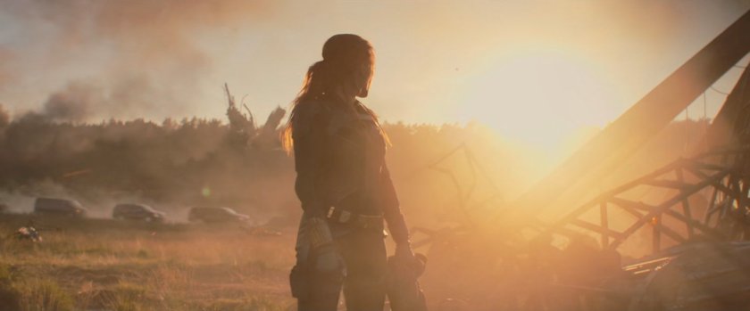 Natasha standing in a field strewn with debris as the sun sets behind her.