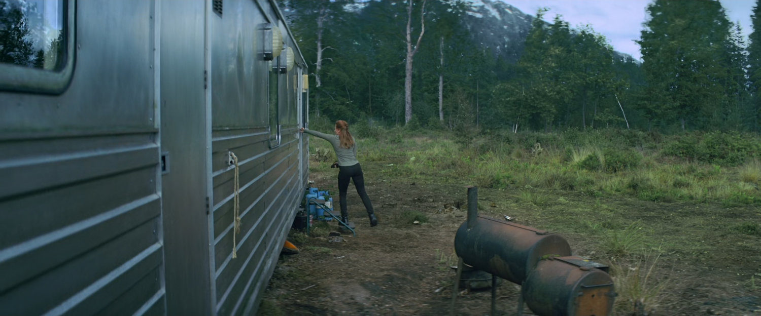 Natasha opening door to mobile home in wooded clearing.