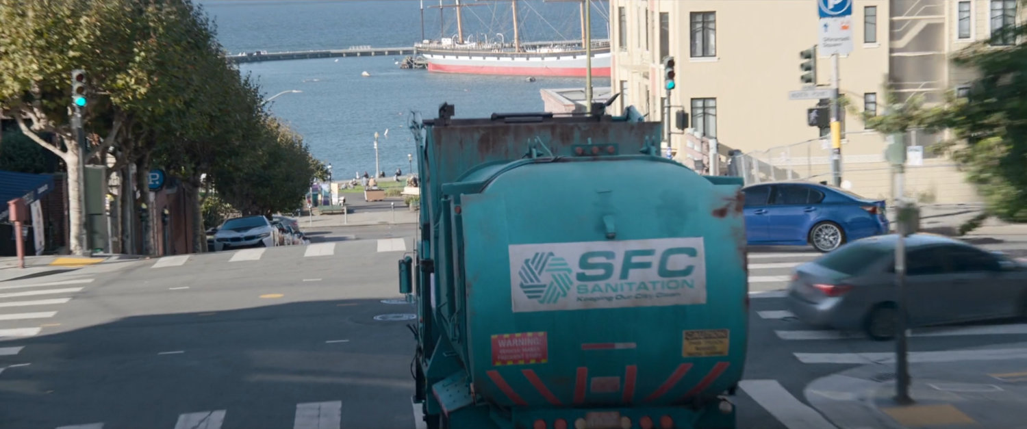 An SFC garbage truck heads down a street with the Bay in background.