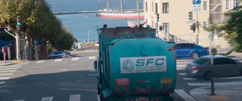 An SFC garbage truck heads down a street with the Bay in background.