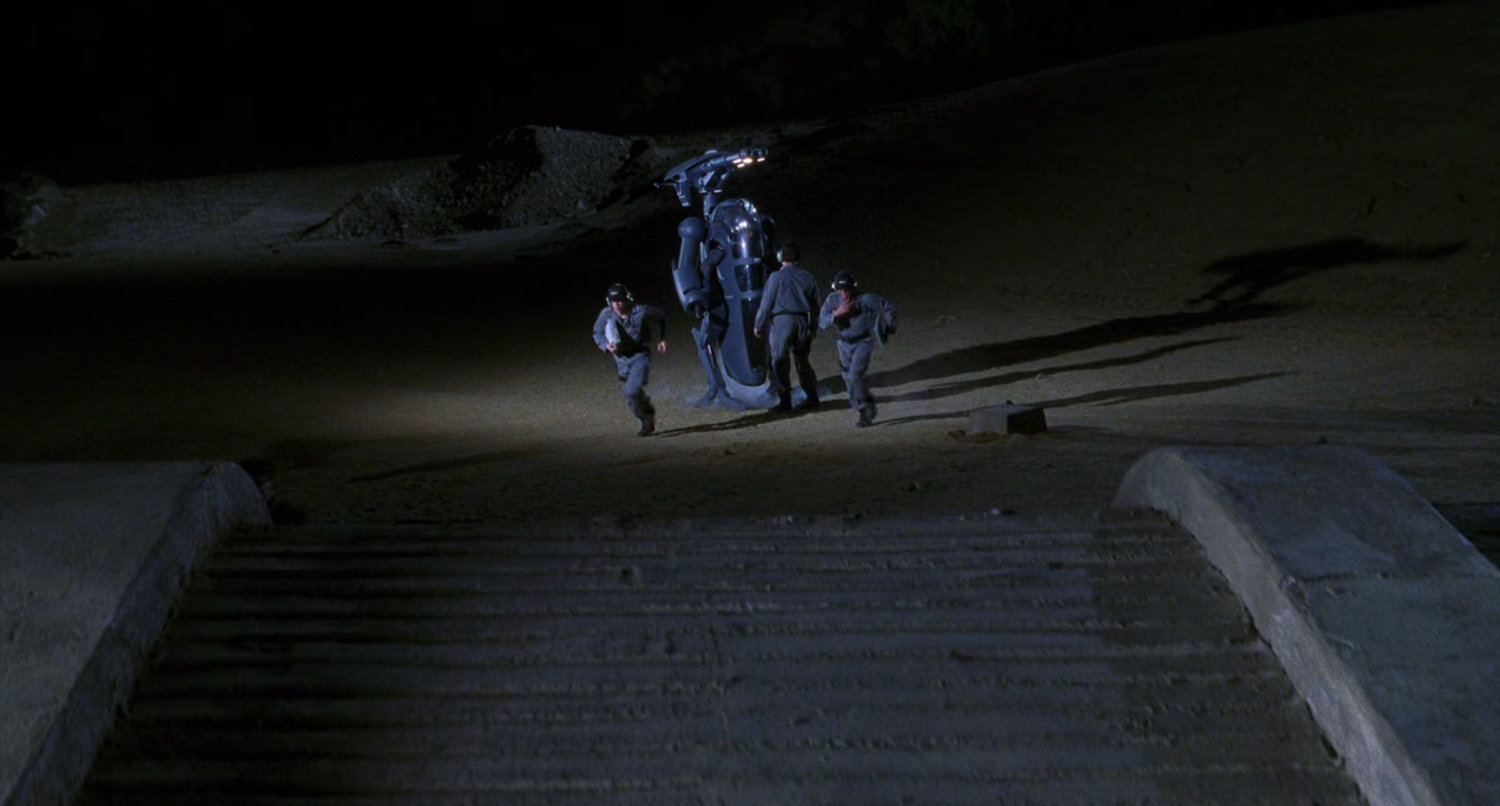 Men setting up robotic suit in desert.