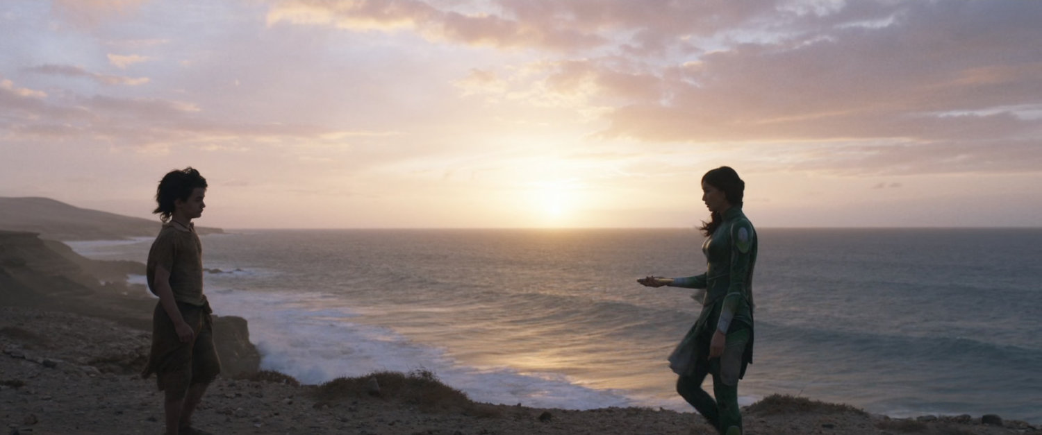 Sersi presents something to a young boy on a cliff overlooking the ocean.