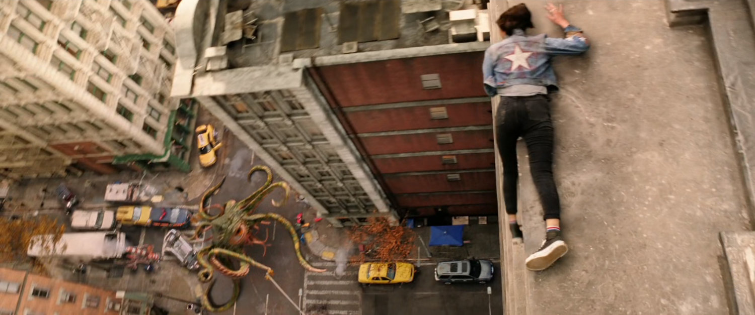 High angle of America Chavez on ledge watching Dr Strange fighting a tentacled beast in an intersection in New York city.
