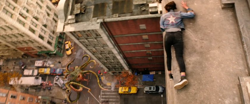 High angle of America Chavez on ledge watching Dr Strange fighting a tentacled beast in an intersection in New York city.