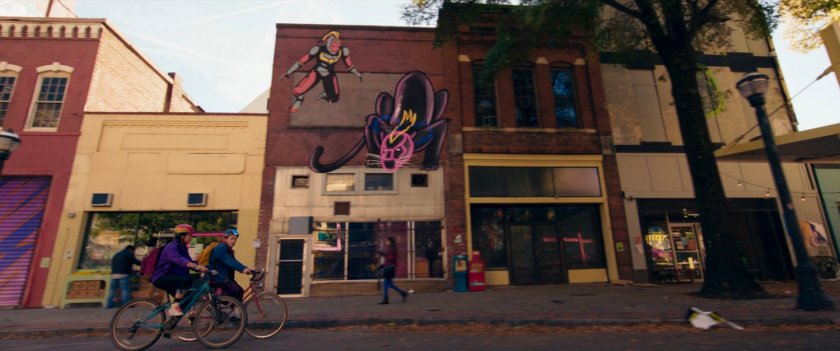 Kamala and Bruno ride bikes past graffiti'd storefronts.