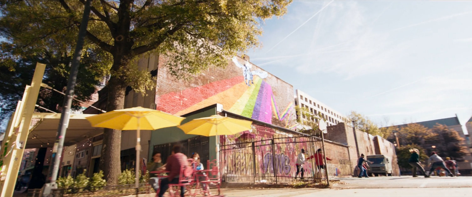 Kamala and Bruno ride bikes past graffiti'd storefronts.