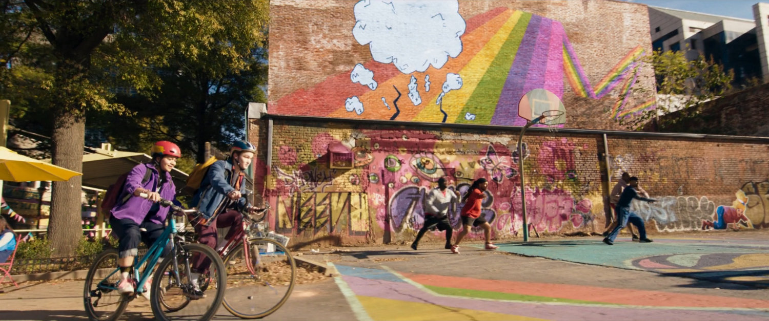 Kamala and Bruno ride bikes past graffiti'd playground.