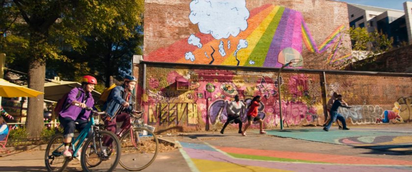Kamala and Bruno ride bikes past graffiti'd playground.