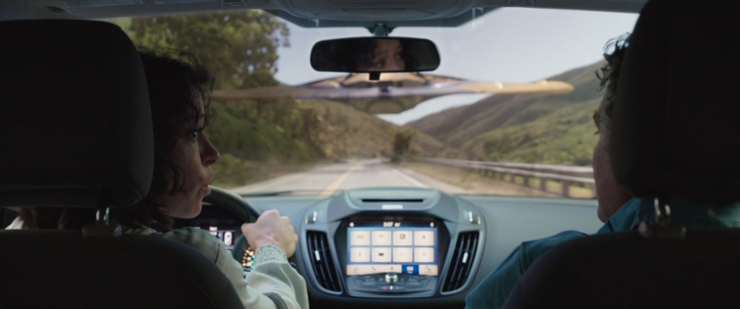 Bruce Banner and Jennifer Walters in a car about to hit a spaceship in the middle of the road.