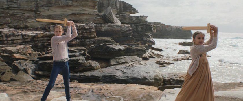 Two girls practice sword poses on the rocks near the ocean.