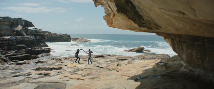 Sif and Axl practice sword fighting on the rocks near the ocean.