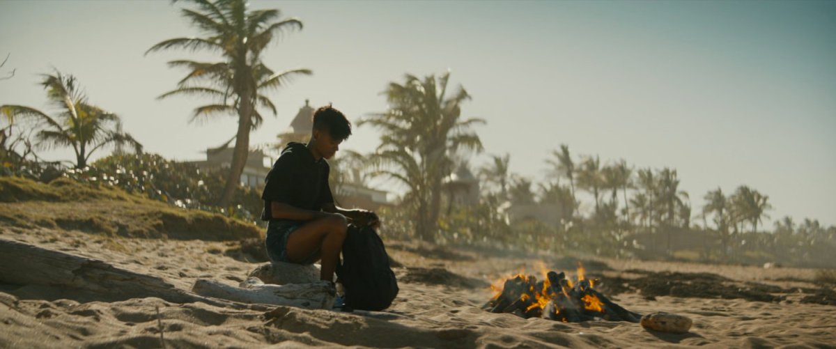 Shuri sits by a campfire on a beach.