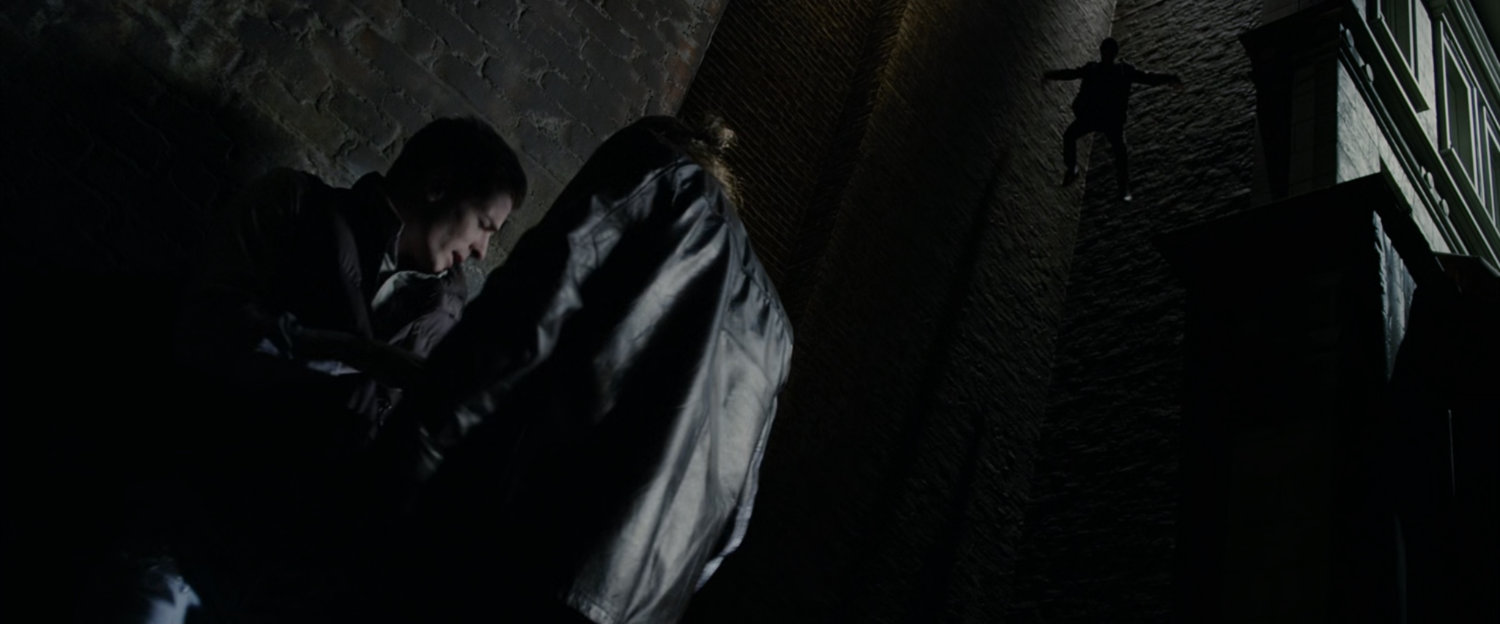 A shadowy figure jumps from a perch on an alley wall towards two thugs.