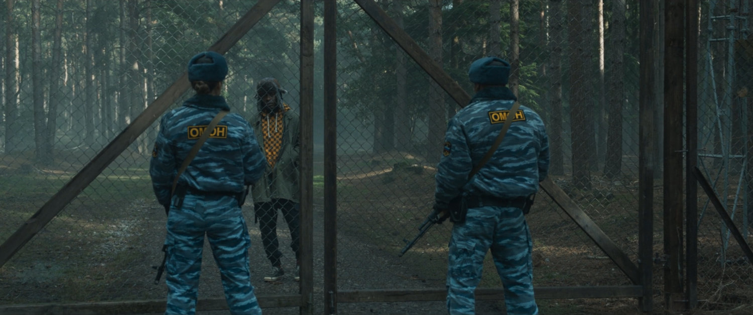 Two guards confront a man by a chain link gate in a misty forest.