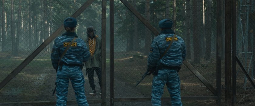 Two guards confront a man by a chainlink gate in a misty forest.